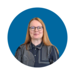 Sinisellä taustalla hymyilevä, punahiuksinen nainen. Naisella on mustasankaiset silmälasit ja mustavalkokuvioinen kauluspaita.