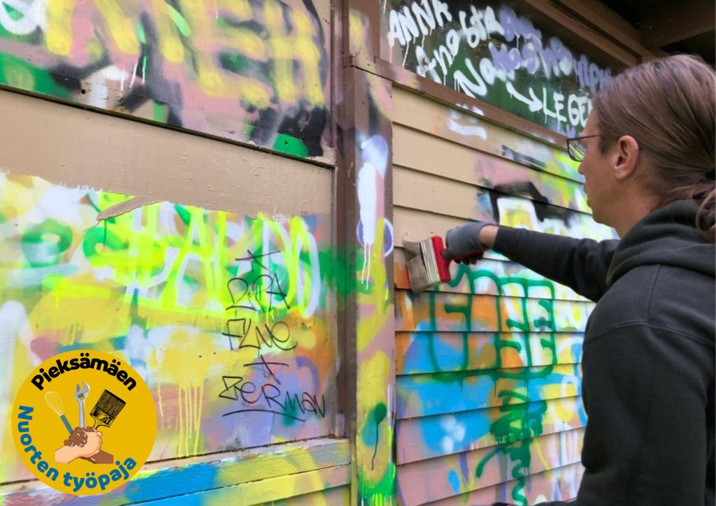 värikästä graffitiseinää korjausmaalataan ruskealla maalilla. Kuvassa henkilö, jolla iso suti kädessä. Alakulmassa keltaisella pohjalla logo, jossa lukee Pieksämäen Nuorten työpaja.