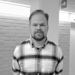 Miesoletettu valkoisessa paidassa jossa mustilla ja harmailla viivoilla ruutuja. Miehellä lyhyet hiukset, viikset ja parta.