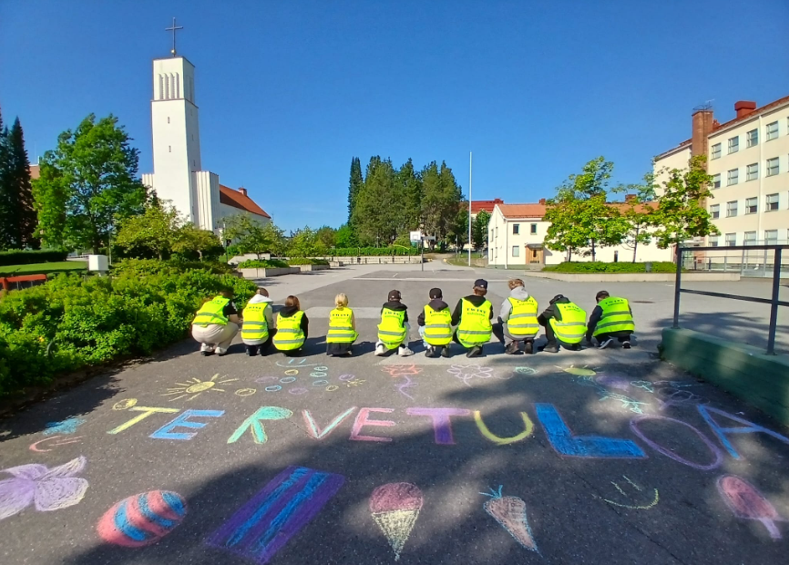 Kesätyönuoria kirkon ja koulun edessä, teksti Tervetuloa kirjoitettu katuun.
