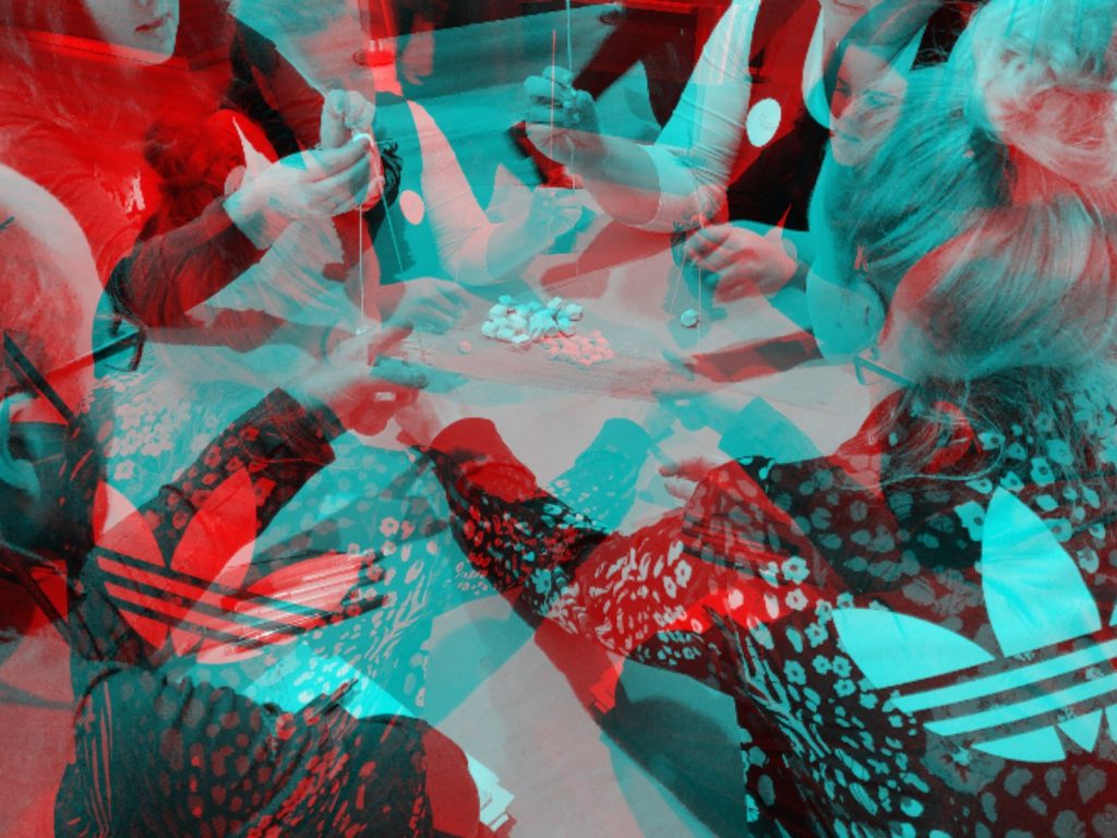 Henkilöitä kokoontunut pyöreän pöydän ääreen tekemään käsitöitä ja suunnittelemassa tulevaa. Kuvassa varjomaisia efektejä punaisen, turkoosin ja mustan sävyissä.