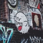 tummalla rähjäisellä graffitiseinällä erilaisia tekstejä ja kuvia sekä spraymaalattu vaalea videopelin virnuileva haamu