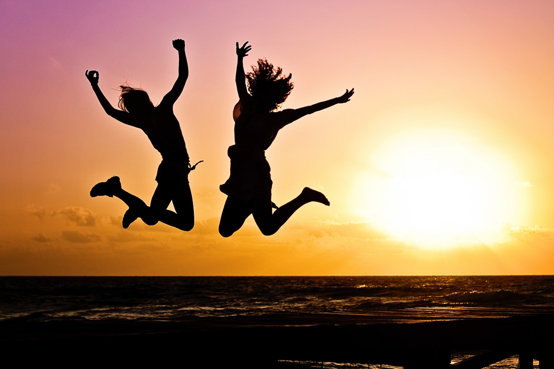 kaksi henkilöä hyppää riemuissaan rannalla keltapinkissä auringonlaskussa