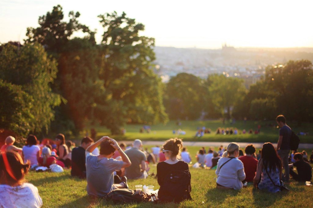 suuri joukko ihmisiä on kokoontunut vehreään puistoon istumaan ja nauttimaan auringosta piknikin merkeissä