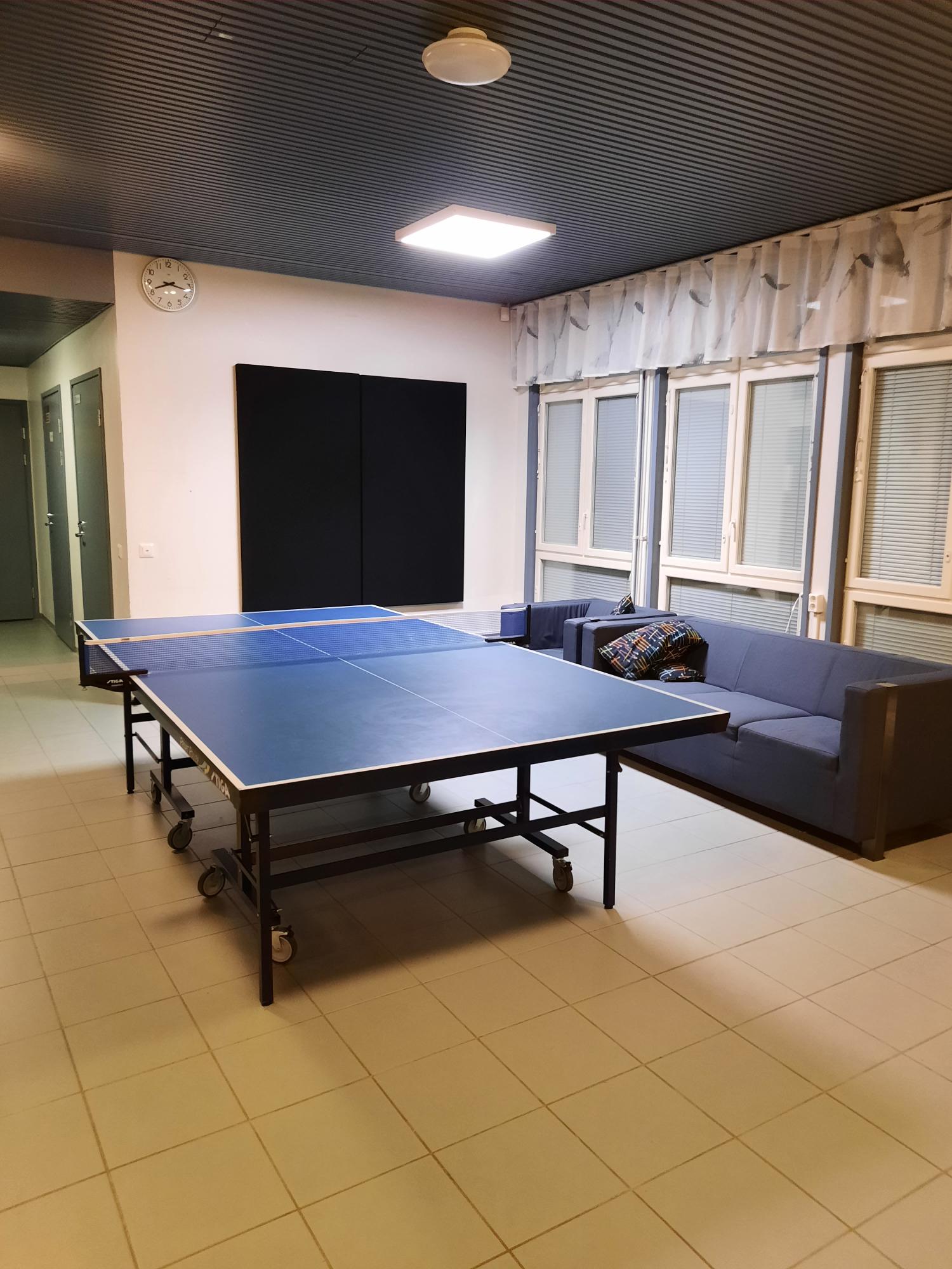 Kuvassa Kurkimäen Pop-up nuokkarin sisätiloista otettu kuva. Tilassa on sininen pingispöytä, siniset sohvat, valkoiset laattalattiat ja valkoiset seinät. Oikealla seinustalla ikkunoita ja vaaleat verhot. Takaseinällä musta neliskanttinen elementti seinällä.