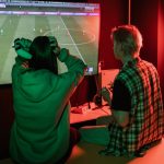 Nuoret pelaavat pleikkarilla jalkapalloa peliä ja nainen pitää käsiään pään päällä kun häntä jännittää