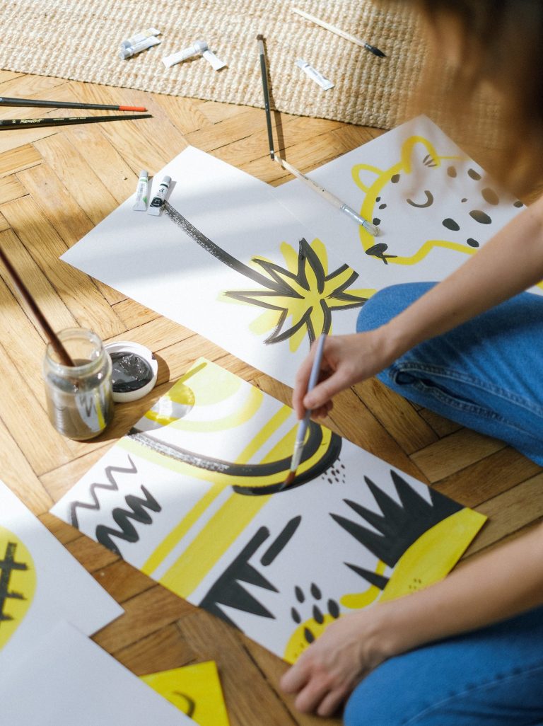 Ihminen maalaa valkoiselle paperille maalausta, jossa mustia ja keltaisia viivoja ja kuvioita.