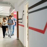 Joukko nuoria kävelee koulun käytävää. Käytävän seinällä on mustia ja punaisia graafisia viivoja. Avainsanoja: koulu, opiskelu, yhteishaku, kaveruus