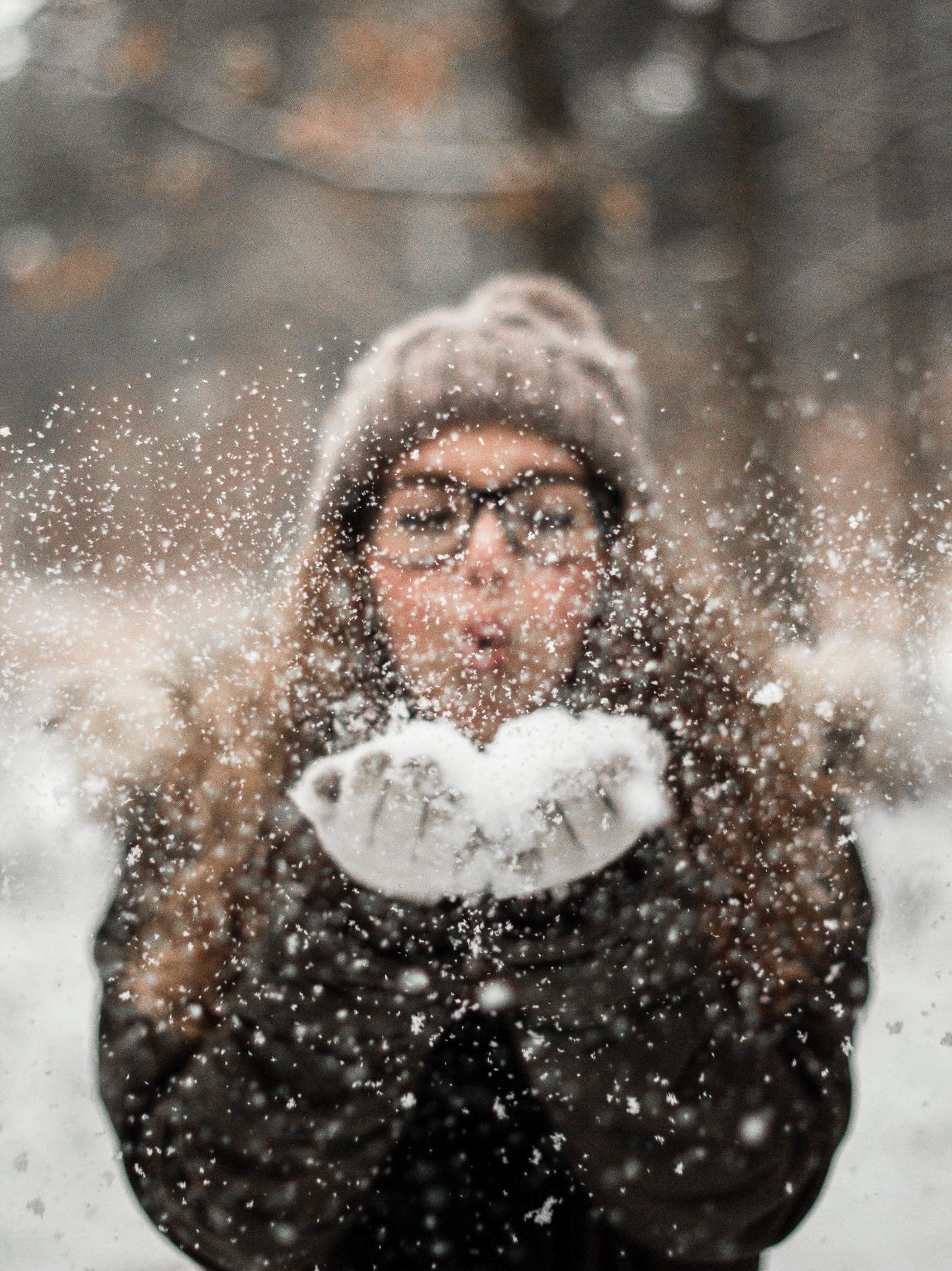 Henkilö puhaltaa lunta käsistään ilmaan. Hänellä on päässään mustasankaiset silmälasit ja ruskea pehmeän näköinen pipo, missä on tupsu. Taustalla näkyy talvisia puita. Avainsanoja: talvi, loma, hiihtoloma, talviloma, ilo, lumi