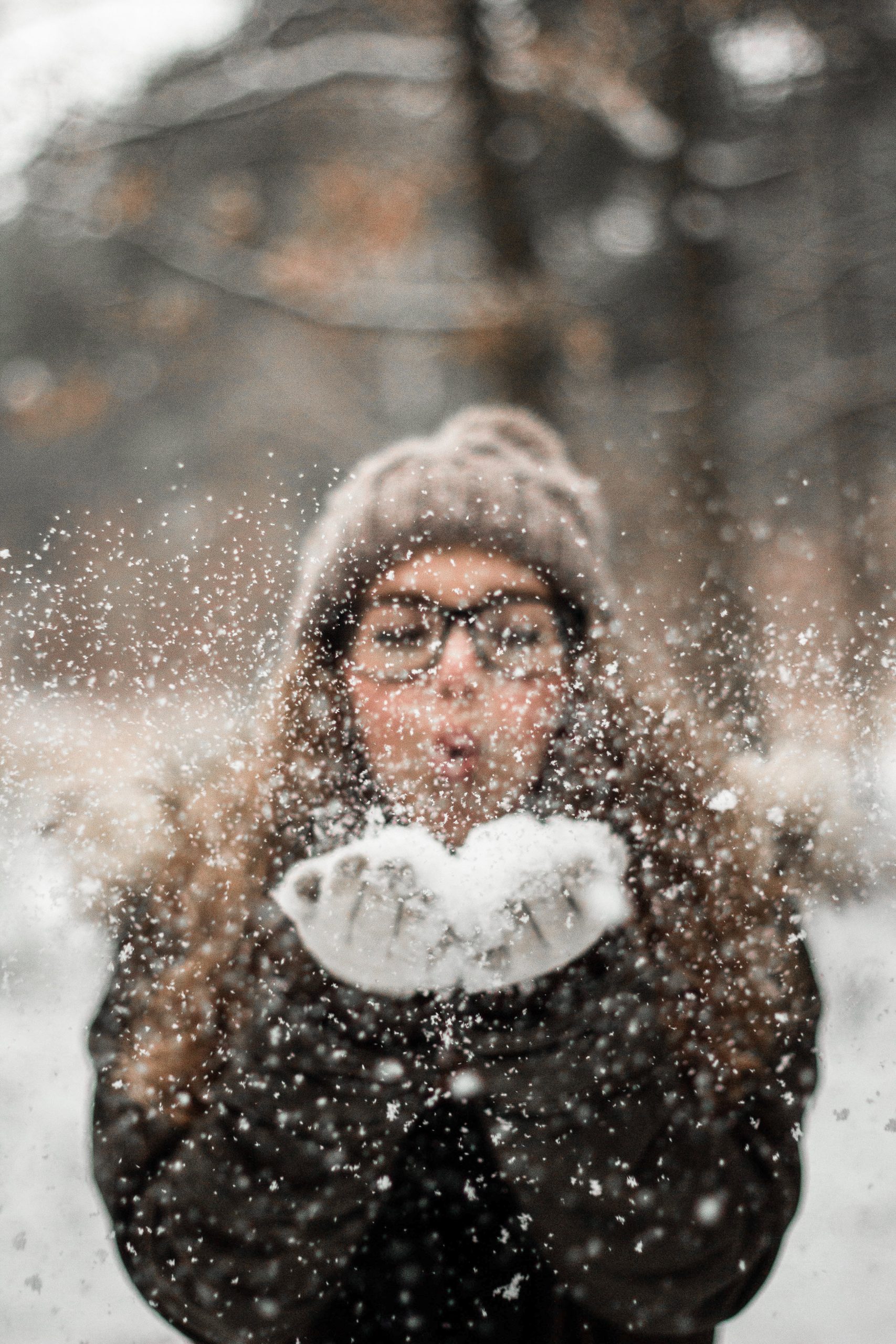 Henkilö puhaltaa lunta käsistään ilmaan. Hänellä on päässään mustasankaiset silmälasit ja ruskea pehmeän näköinen pipo, missä on tupsu. Taustalla näkyy talvisia puita. Avainsanoja: talvi, loma, hiihtoloma, talviloma, ilo, lumi
