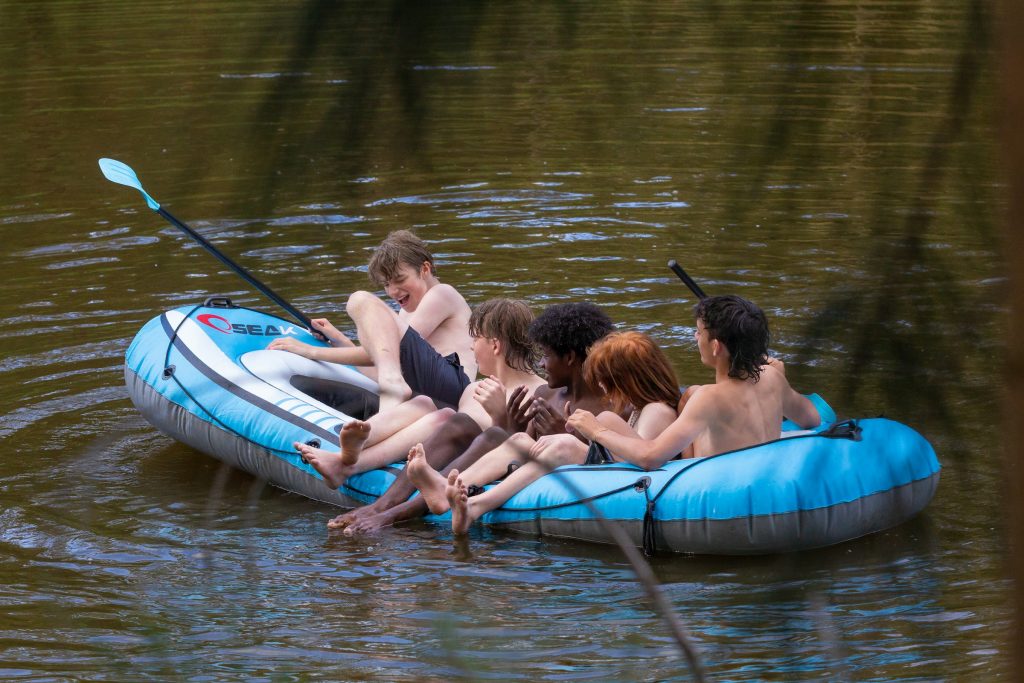 Viisi nuorta istuu sinisessä kumiveneessä järvessä nauraen ja iloiten. Kahdella nuorista on käsissään mela. Avainsanoja: kesä, loma, kesäloma, ilo, vapaa-aika, kaveruus
