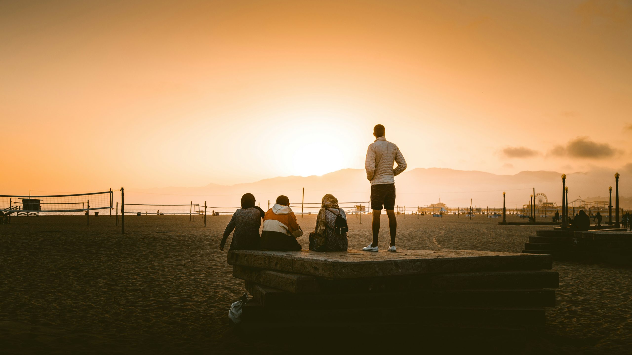 Neljä ihmistä katsoo kohti laskevaa aurinkoa hiekkarannalla. Taustalla näkyy rantalentopalloverkkoja ja huvipuistolaitteita. Kuvassa on lähes seepian värinen sävytys.