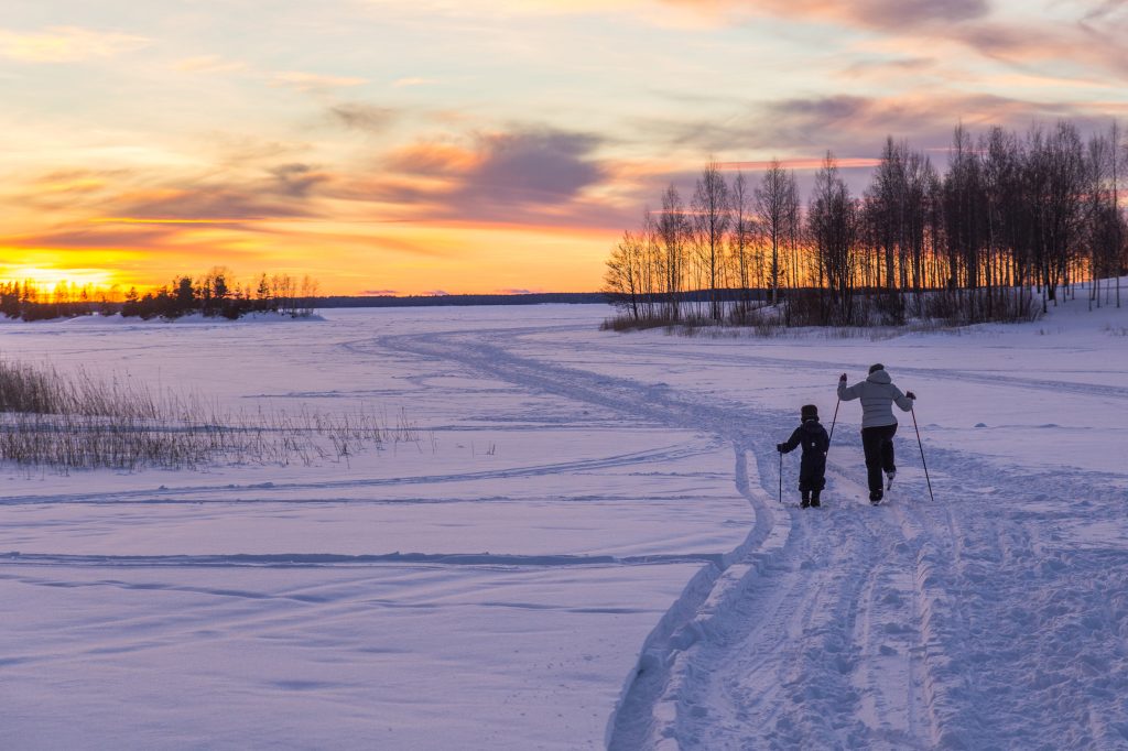 Aikuinen ja lapsi hiihtämässä ladulla järven rannalla keltaisen auringon laskiessa. Avainsanoja: hiihtoloma, loma, hiihto, talvi, ilo, liikunta, luonto