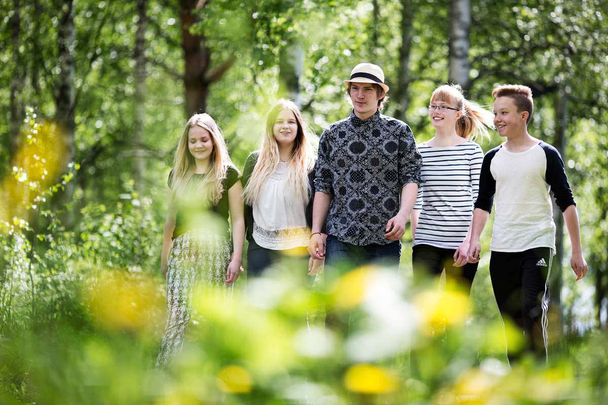 Viisi nuorta kävelee hymyillen kohti kameraa kesäisessä puistossa. Kuva on otettu vihreiden puiden lehtien takaa ja se on tarkennettu kasvoihin. Avainsanoja: ilo, kesä, kesäloma, vapaa-aika, kaveruus, luonto, ympäristö, retki