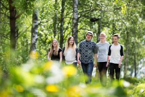 Viisi nuorta kävelee hymyillen kohti kameraa kesäisessä puistossa. Kuva on otettu vihreiden puiden lehtien takaa ja se on tarkennettu kasvoihin. Avainsanoja: ilo, kesä, kesäloma, vapaa-aika, kaveruus, luonto, ympäristö, retki