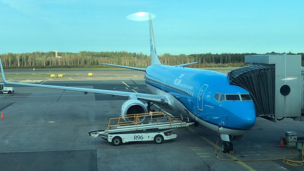 Sininen lentokone lentokentän terminaaliputkessa. Vieressä porrasauto ja taustalla puita sekä sininen taivas.