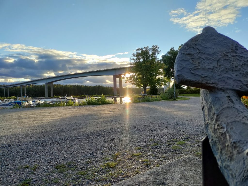 Kuvassa näkyy kauempana Puumalan silta jonka taakse aurinko on laskeutumassa. Oikeassa kulmassa kuvaa näkyy ankkuri ja sen takana hieman kauempana näkyy tammi