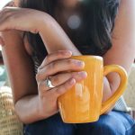 Nuori nainen pitää keltaista kahvikuppia kädessään nojaten polviinsa. Naisella on pitkät ruskeat hiukset, siniset farkut ja sormus oikean käden etusormessa.