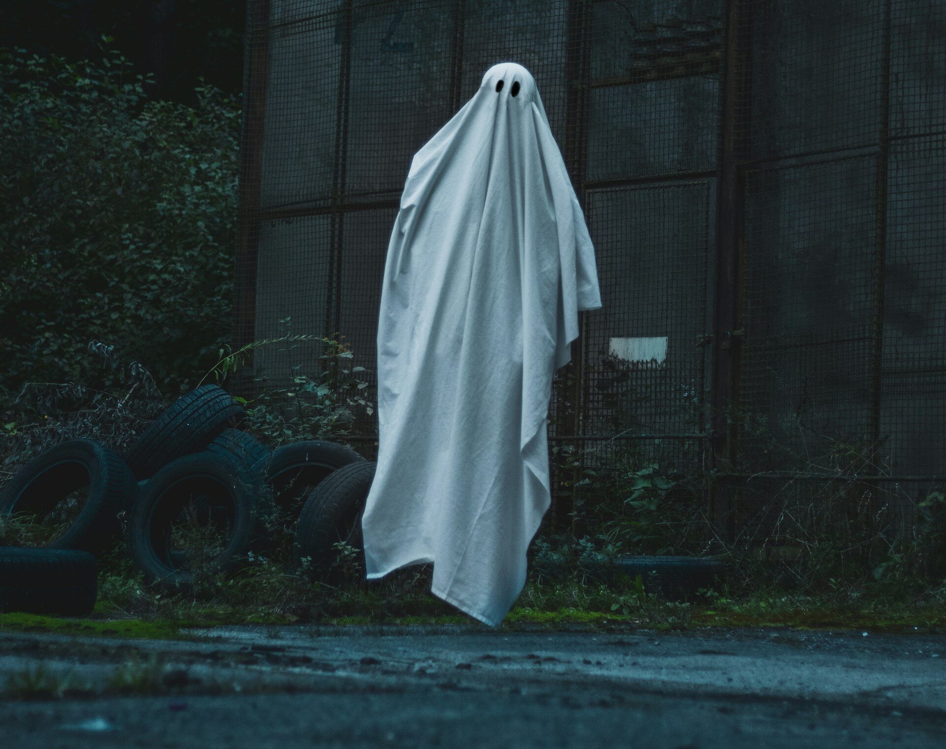 Valkoinen lakanasta tehty kummitus seisoo aavemaisesti häkkirakennelman ja vanhojen auton renkaiden edustalla. Kuva on tumma ja pimeähkö.