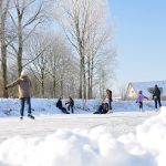 Nuoret luistelevat ja leikkivät luonnonjäällä aurinkoisessa talvisäässä. Taustalla näkyy puita ja taloja