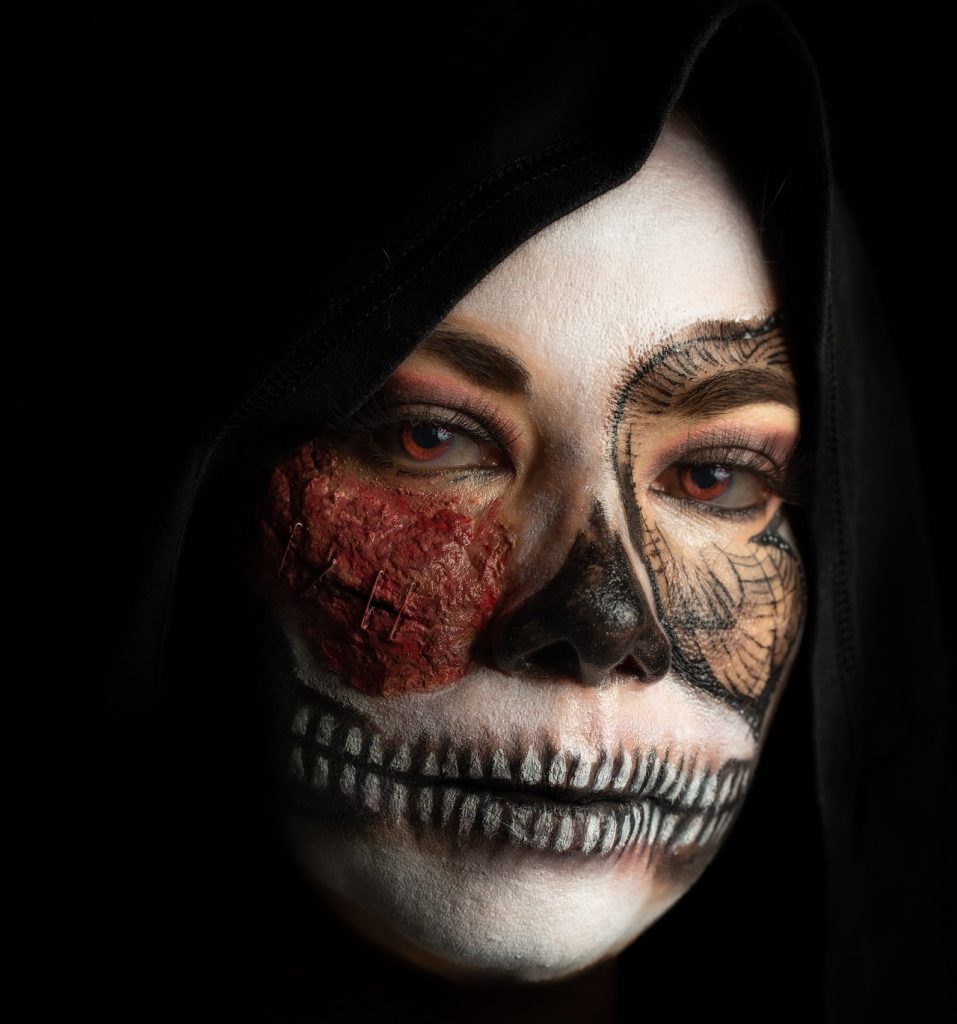 Lähikuva naisen kasvoista, jotka ovat maalattu pääkallon mallisiksi. Naisen päässä huppu, joka kehystää kasvoja.