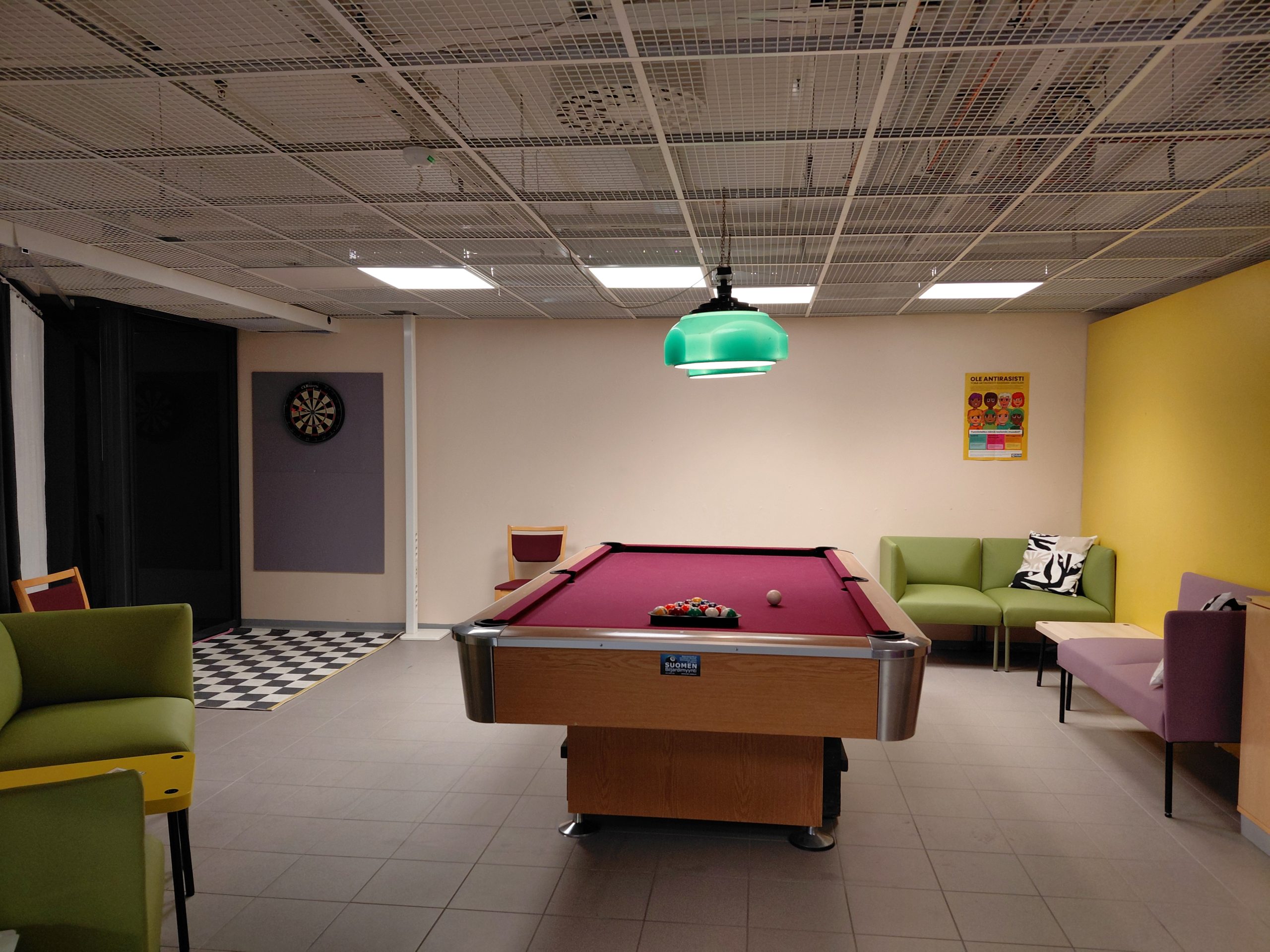 Vasemmalla on tummat verkot, jotka estävät käytävältä tilaan katsomisen. Takaseinän vasemmalla reunalla on darts-taulu, joka takana on suojalevy. Heittoalueella on mustavalkoinen shakkikuvioitu muovimatto. Keskellä tilaa on tummanpunaisella verkakankaalla oleva biljardipöytä. Vasemmalla näkyy biljardipöydän vasemman etukulman luona vihreä sohvaryhmä sohvapöydän kanssa. Takaseinällä oikealla on vihreä sohva, jonka edessä keltaista väliseinää vasten sohvapöytä. Sohvapöydän vieressä seinää vasten on punertava sohva.