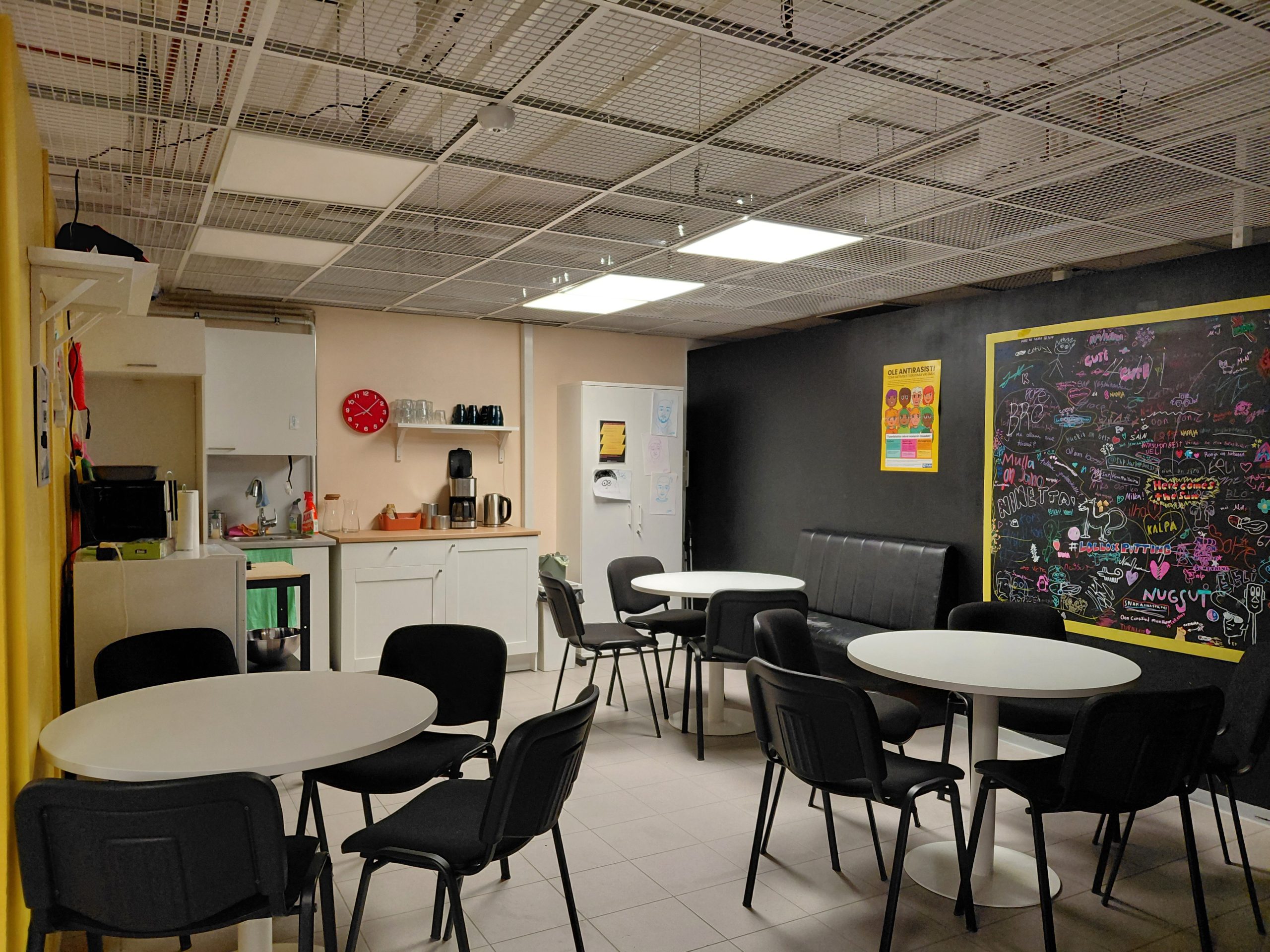 Näkymä minikeittiöön, jossa takaseinällä on jääkaappi ja pieni keittiötila. Seinällä on punainen kello hyllyn vieressä, jossa säilytettään mukeja ja laseja. Vasempaan väliseinään on kiinnitetty hylly, jonka alapuolella on mikro ja pieni jääkaappi. Tilan täyttää kolme valkoista pyöreää pöytää, joiden ympärillä on mustia kangaspäällysteisiä tuoleja. Kauimman pöytäryhmän luona on myös pieni musta sohva seinää vasten. Oikean puoleinen seinä on musta ja seinästä on rajattu keltaisin raamein osio, jossa on kirjoitusta.