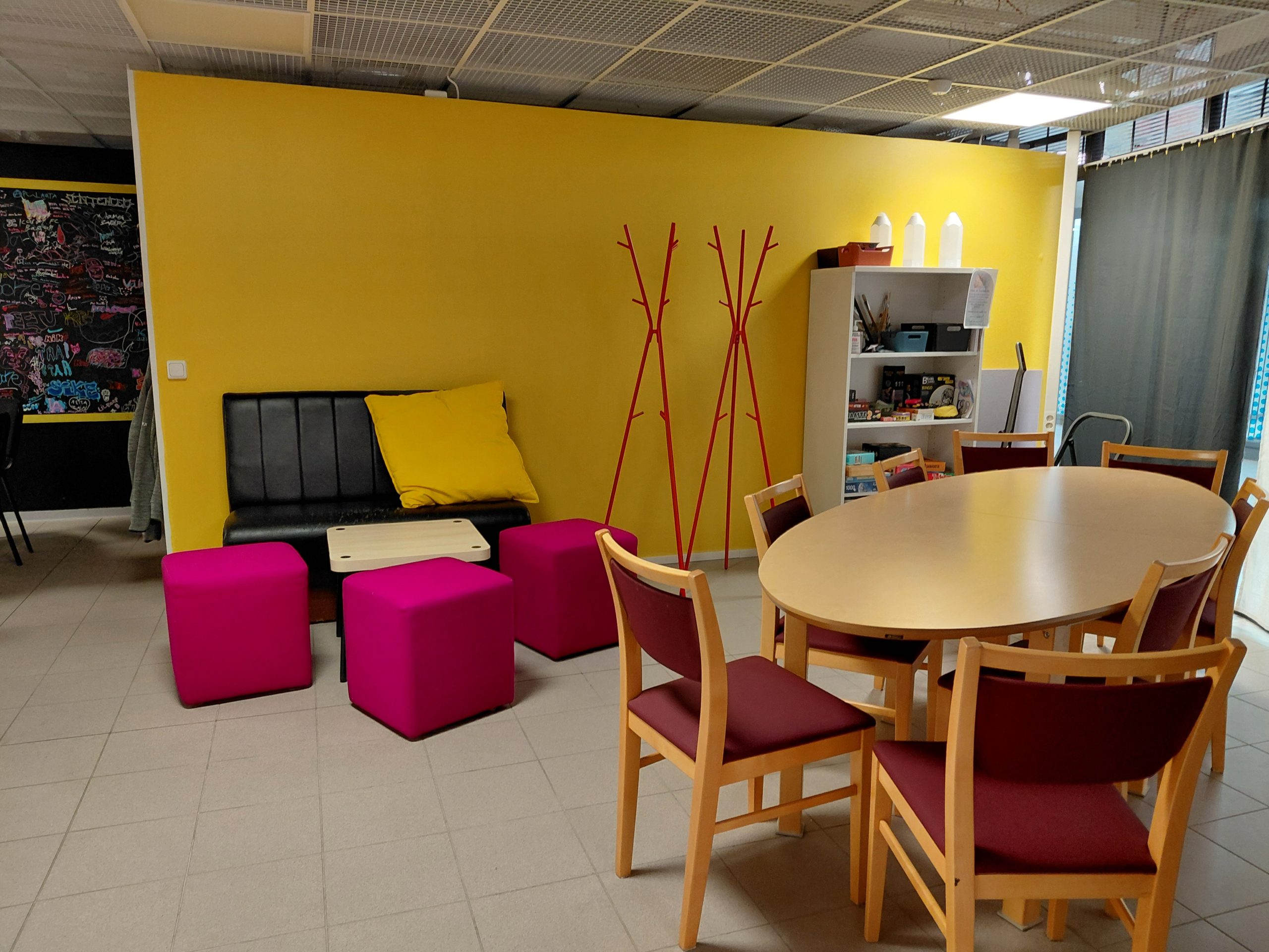 Näkymä Napajan tilaan. Tilan takaosassa kirkkaankeltainen seinäke, jonka takaa näkyy musta takaseinä, jossa kirjoitusta keltaisten raamien sisällä. Keltaisen seinän edessä pieni musta sohva, jolla keltainen iso tyyny Sohvan edessä pieni kahvipöytä, joka ympärillä kolme pinkkiä rahia. Sohvan oikealla puolella kaksi punaista kolmijalkaista naulakkoa tyhjillään. Naulakoiden oikealla puolella valkea avohyllykkö, jossa lautapelejä ja askartelutarvikkeita. Oikealla näkyy harmaa verho, joka estää näkymän kauppakeskuksen käytävälle lasiseinä läpi. Etualalla on vielä ovaali puupöytä, jonka ympärillä on kahdeksan puupuolia tummanpunaisilla verhoiluilla.