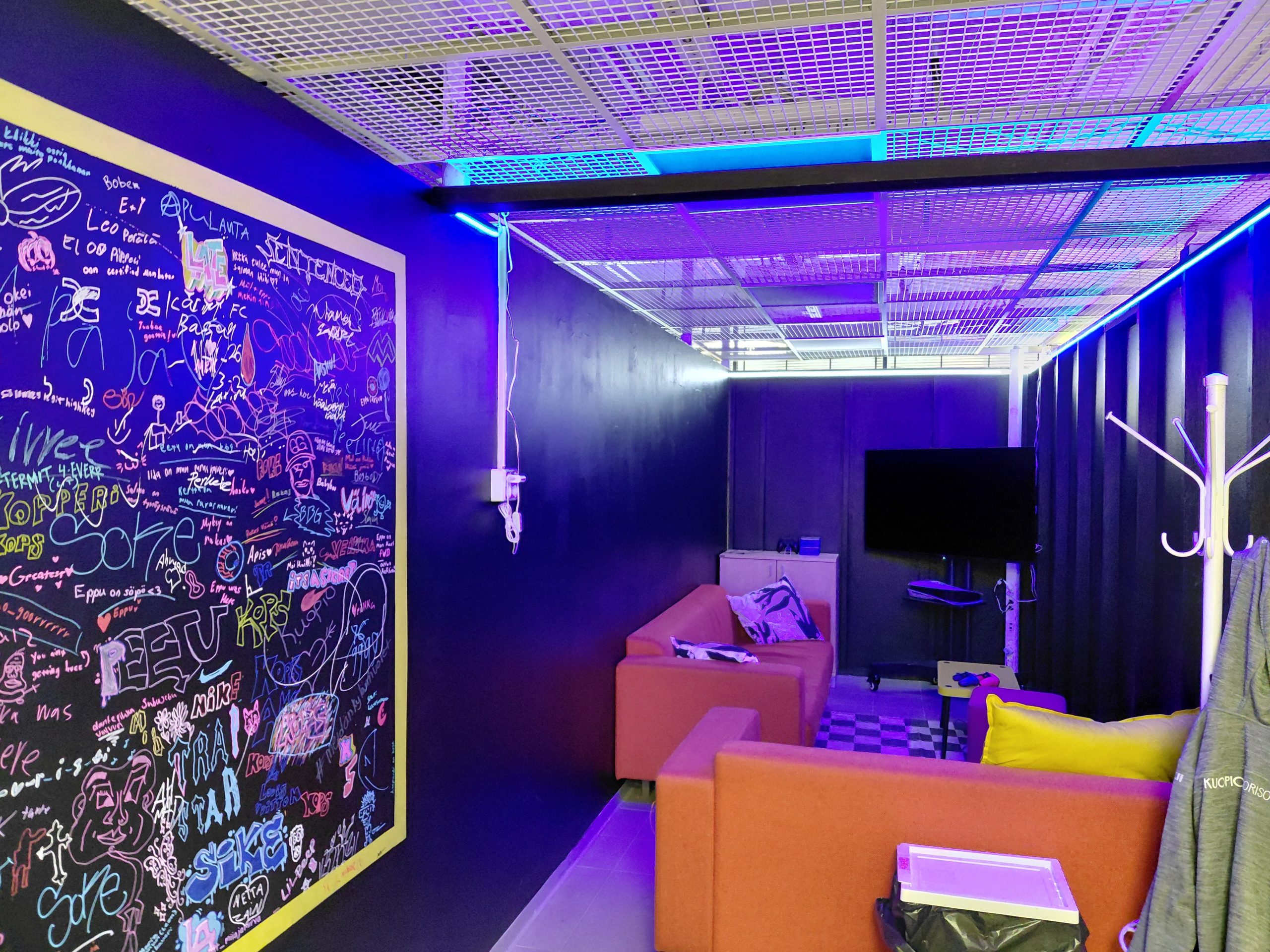 UV-valolla valaistu pelinurkkaus. Kaiki seinät ovat maalattu mustiksi ja vasemmalla seinällä on kirjoitusta, joka hohtaa UV-valossa. Kirjoitukset ovat keltaisten raamien sisällä. Nurkkauksen takaseinän luona on pieni kaapisto, jonka edessä oranssi sohva. Takaseinän luona on myös iso televisio ja PS5-konsoli telineellä. Television ja sohvan luona on myös pieni pöytä. Toinen sohva rajaa nurkkauksen erilleen muusta tilasta olemalla poikittain oikean seinän vierellä. Sohvan takana on valkoinen naulakko.