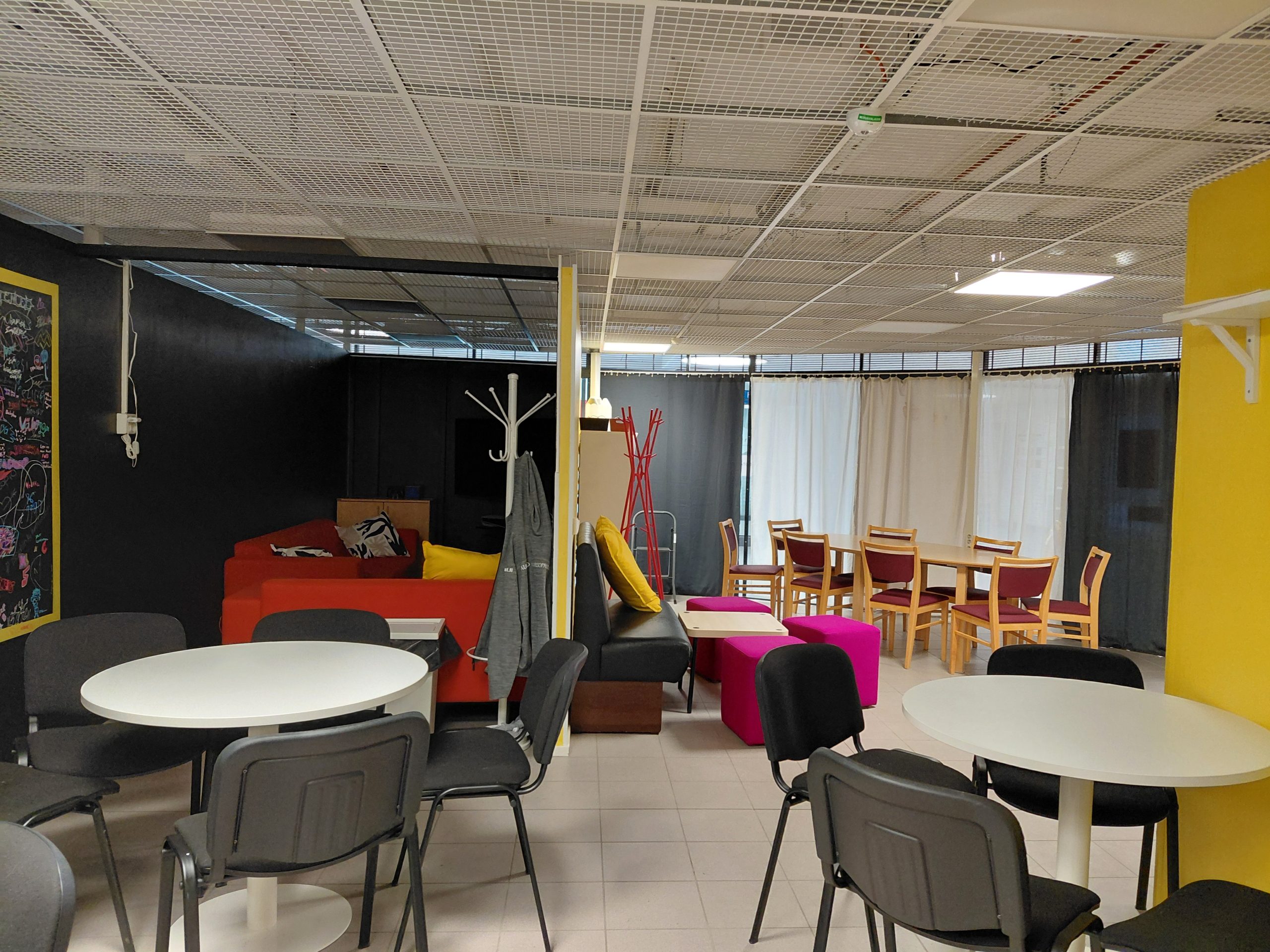 Minikeittiön suunnasta näkymä tilaan. Vasemmalle jää pelinurkkaus oikealla puolella näkyy tilan sisääntulo ja istuinryhmät. Minikeittiön puolella on valkeita pyöreitä pöytiä, joiden ympärillä on mustia kangaspäällysteisiä tuoleja.