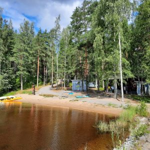 Kuvassa kesäinen uimaranta, jossa rannalla suppilauta ja soutuvene. Puiden takana mökki.