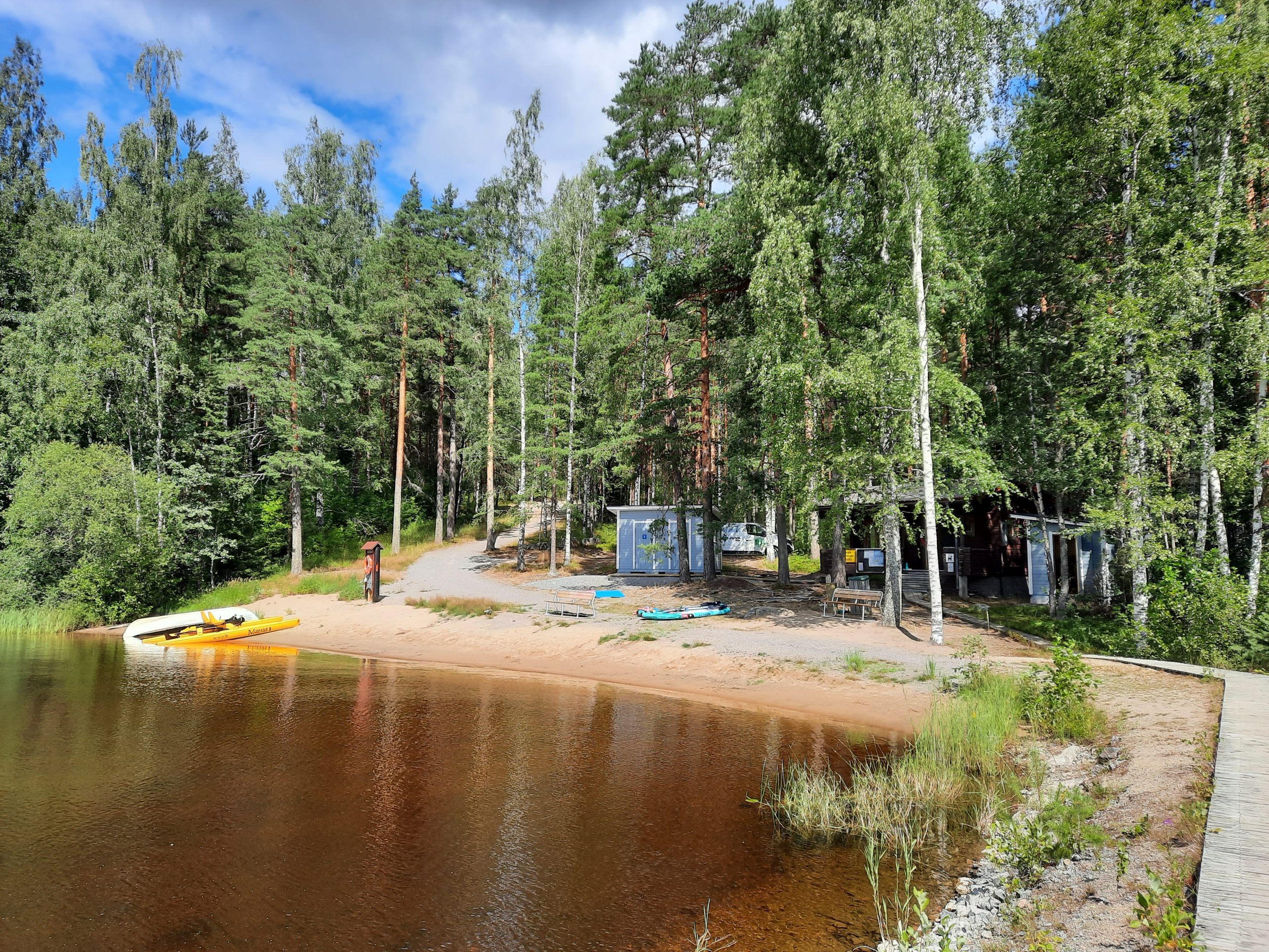 Kuvassa kesäinen uimaranta, jossa rannalla suppilauta ja soutuvene. Puiden takana mökki.